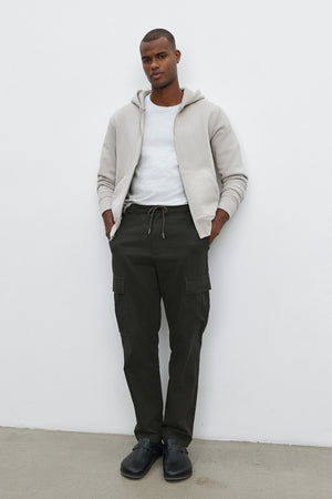 A man stands against a white wall wearing the REN HOODIE by Velvet by Graham & Spencer, a white t-shirt, dark green cargo pants, and black shoes, with his hands in his pockets.