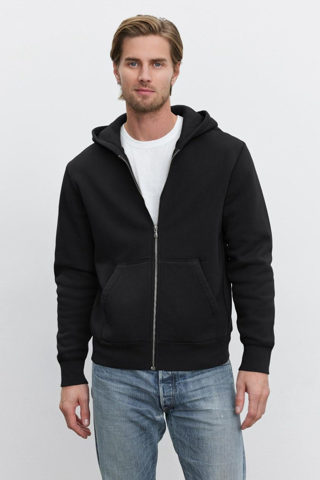   A man wearing a black REN HOODIE by Velvet by Graham & Spencer over a white t-shirt and blue jeans stands against a plain white background, giving off a subtle vintage look. 