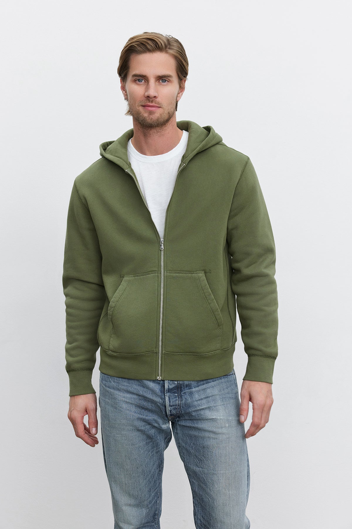   A man with short hair is standing against a plain background, wearing a white T-shirt, blue jeans, and an olive green REN HOODIE with a vintage look from Velvet by Graham & Spencer. 