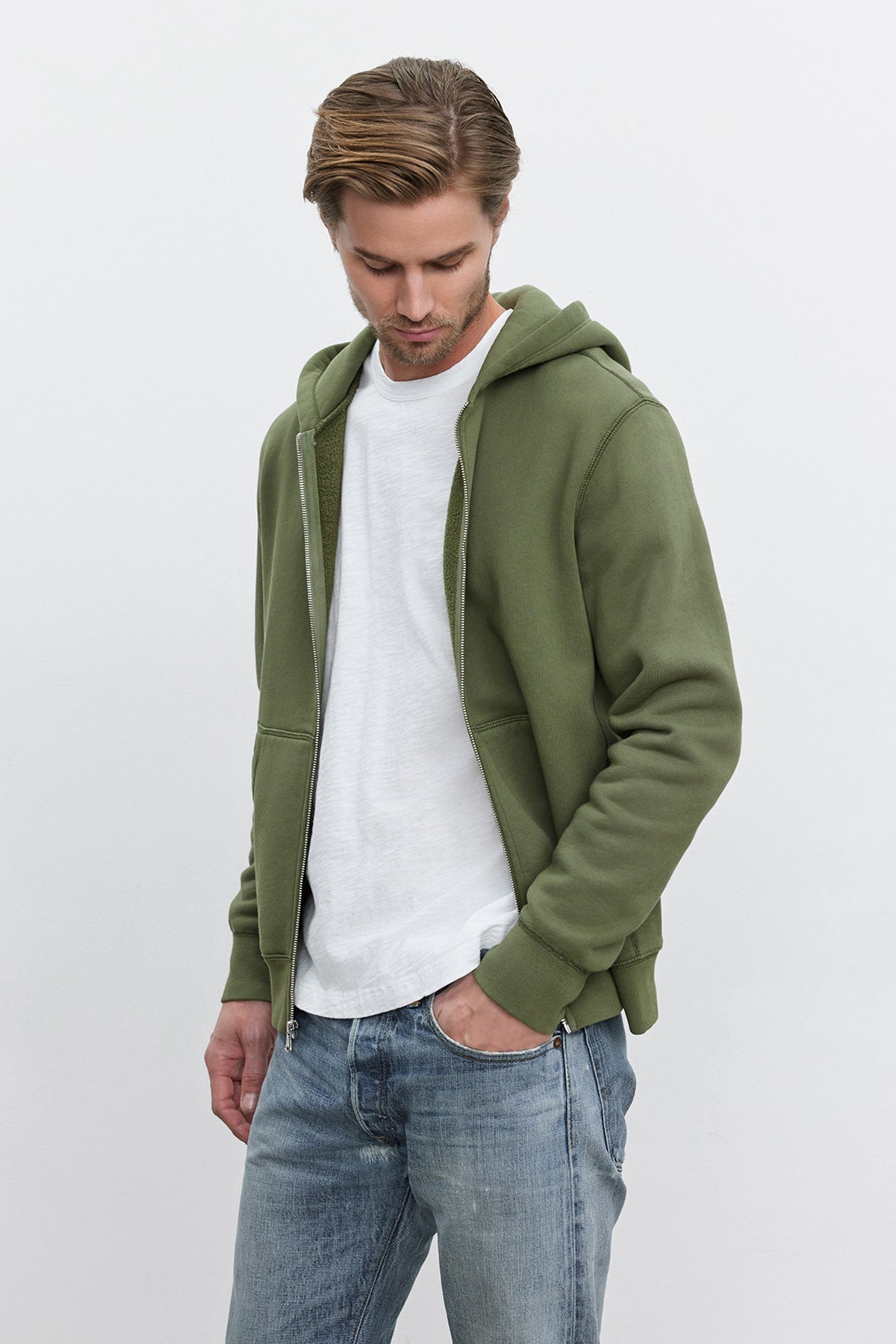   A man wearing the REN HOODIE from Velvet by Graham & Spencer, a green full-zip cotton fleece hoodie, over a white T-shirt and blue jeans stands against a plain background, looking down and exuding a vintage look. 