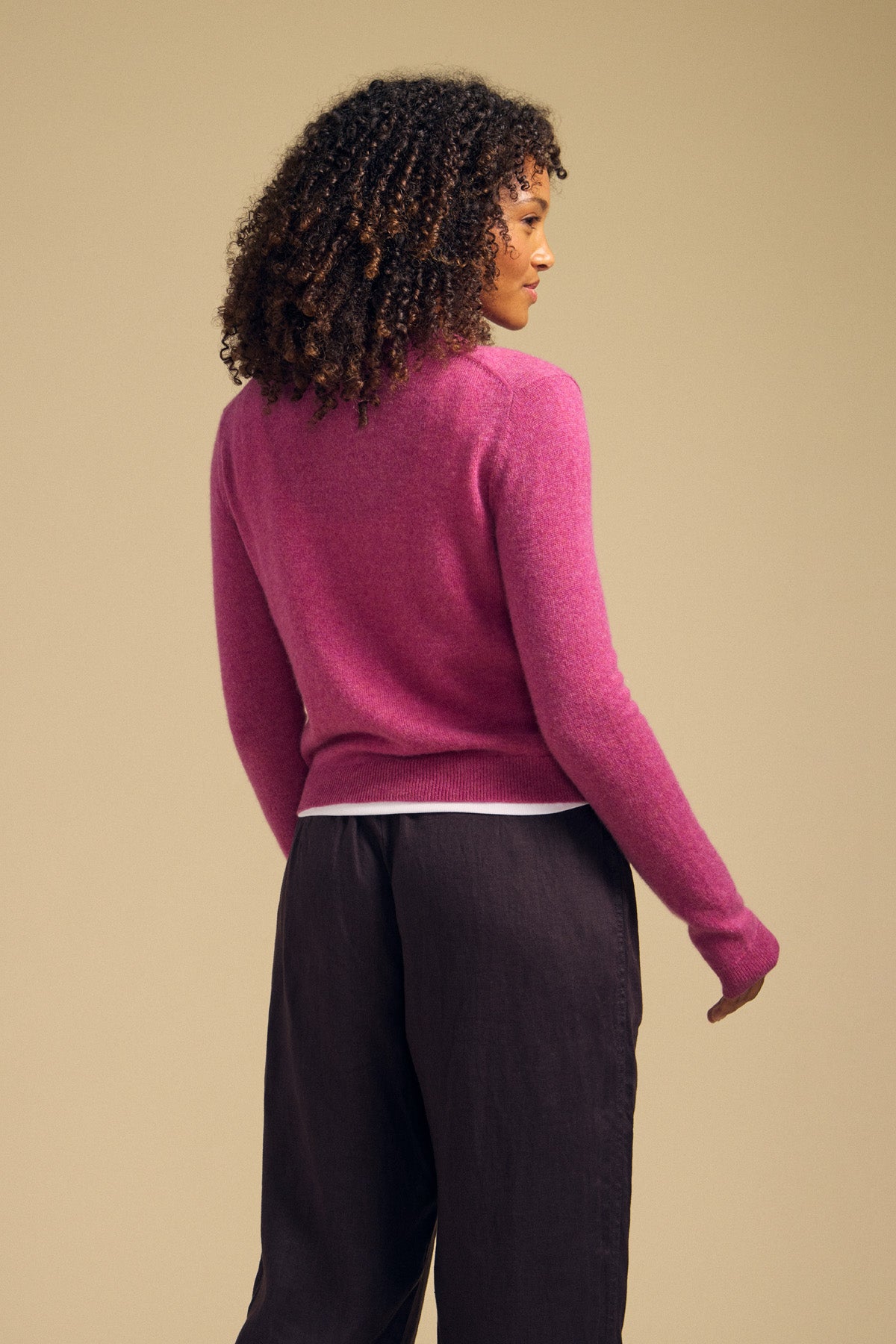 A person with curly hair stands facing away, wearing the AYLINA CASHMERE CARDIGAN by Velvet by Graham & Spencer in pink and dark pants against a neutral background.-42063717073089