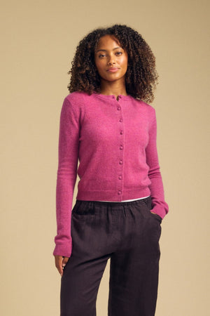 A person wearing the AYLINA CASHMERE CARDIGAN by Velvet by Graham & Spencer in bright pink and dark pants stands with one hand in their pocket, facing forward against a beige background.