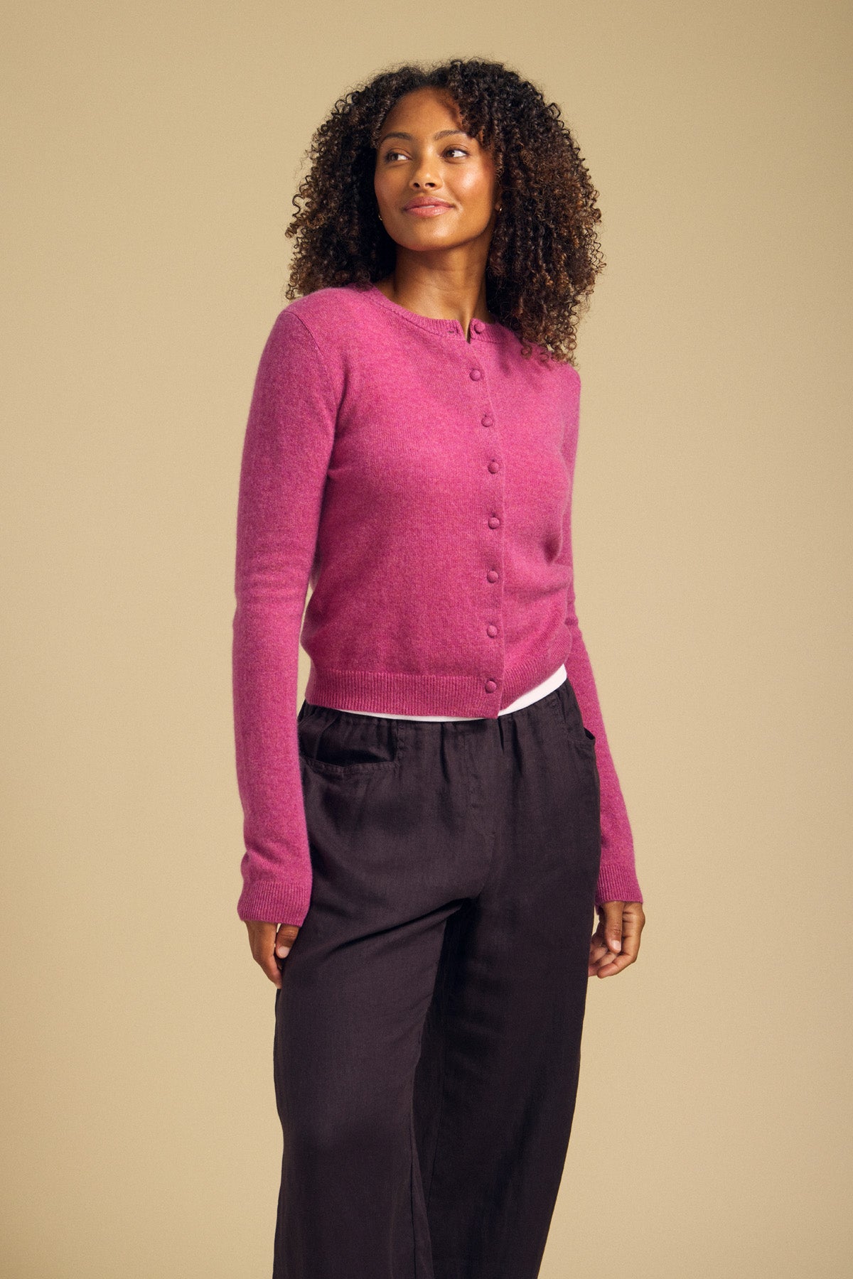 A woman stands against a beige background, wearing the AYLINA CASHMERE CARDIGAN by Velvet by Graham & Spencer—a cropped pink sweater layered over a white shirt and loose black pants for effortless style.