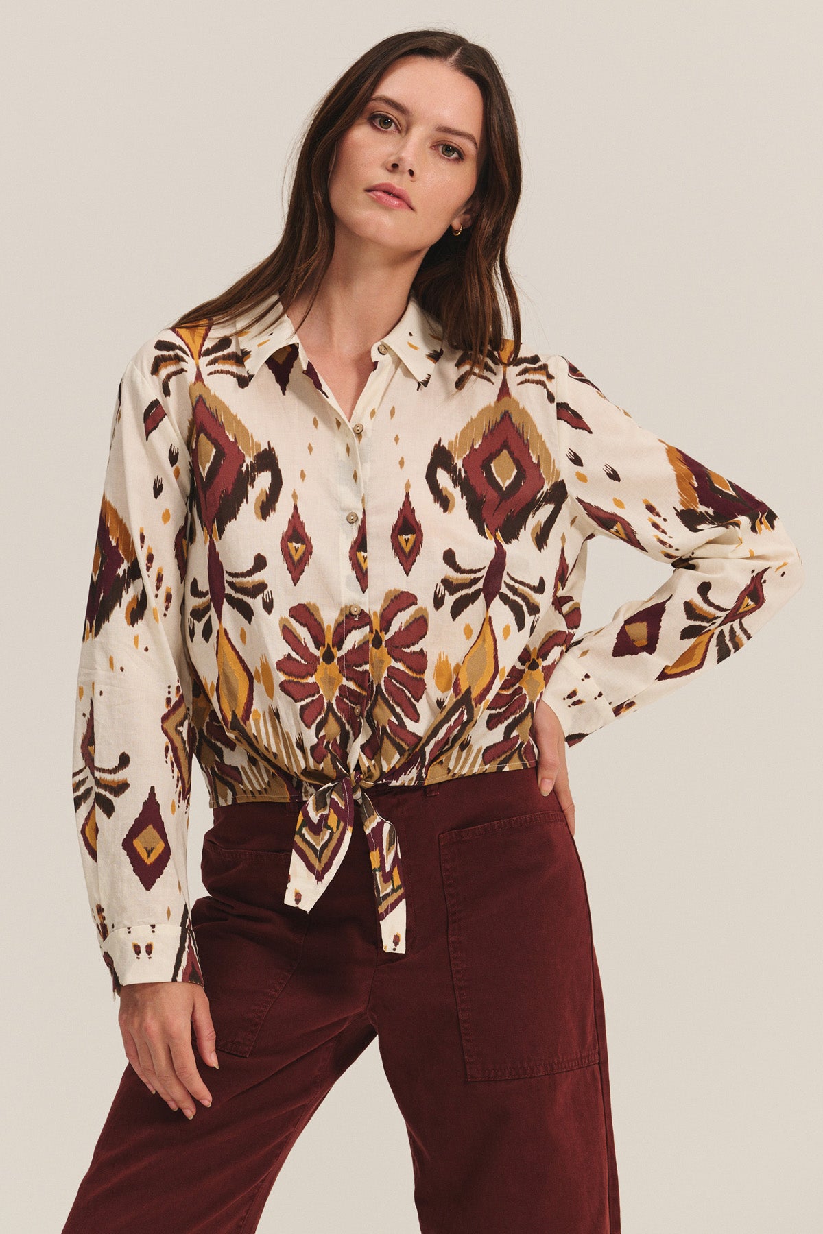 Wearing the KAYLA TOP by Velvet by Graham & Spencer—a cropped button-down blouse with a front tie and long sleeves in a soft cotton-viscose blend—paired with high-waisted burgundy pants against a plain background.-40681722872001