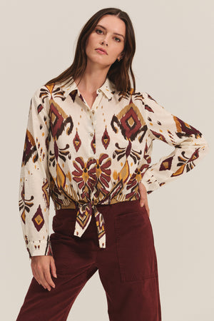 Wearing the KAYLA TOP by Velvet by Graham & Spencer—a cropped button-down blouse with a front tie and long sleeves in a soft cotton-viscose blend—paired with high-waisted burgundy pants against a plain background.