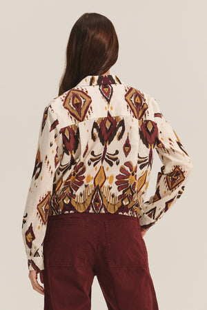A person with long brown hair is shown from the back wearing the KAYLA TOP by Velvet by Graham & Spencer, a cotton-viscose blend button-down featuring geometric and abstract designs, paired with burgundy pants.