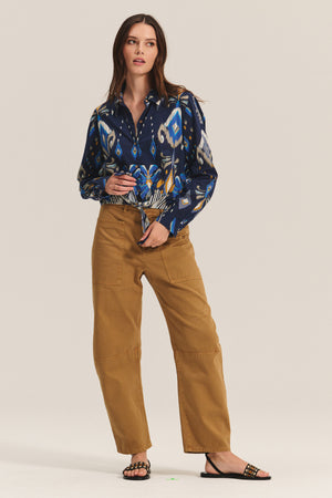 A woman wears the KAYLA TOP by Velvet by Graham & Spencer—a blue patterned button-down cotton-viscose blend shirt—paired with tan high-waisted patch-pocket pants and black sandals, standing against a plain background.