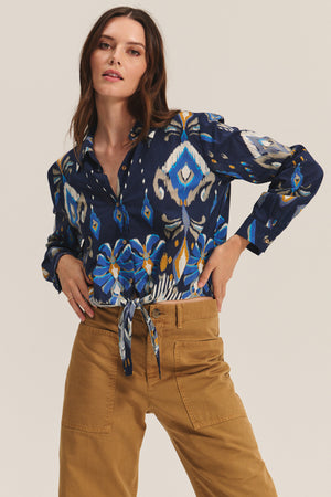 A woman wears the KAYLA TOP by Velvet by Graham & Spencer—a blue patterned cropped button-down blouse with a front tie—paired with tan high-waisted pants, standing hands on hips against a plain background.