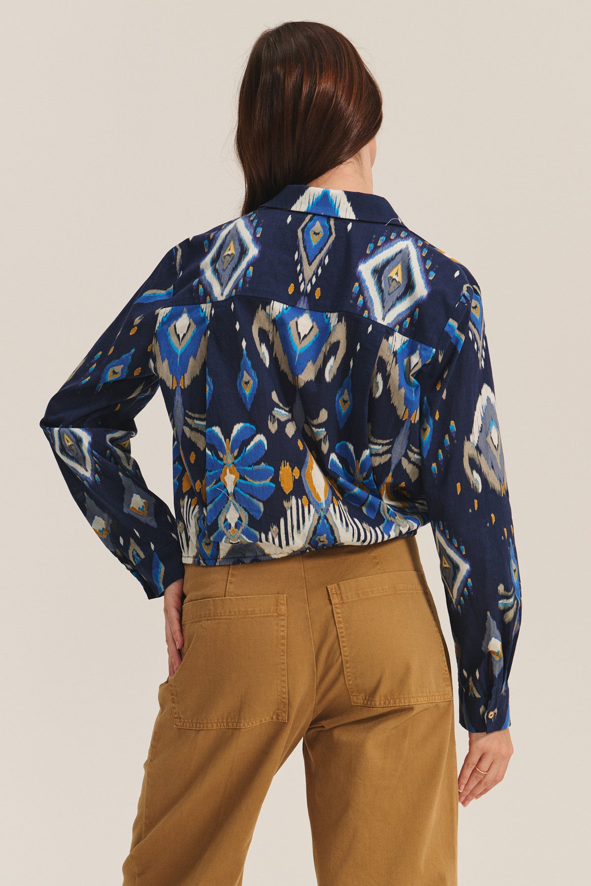 A person stands facing away, wearing the KAYLA TOP by Velvet by Graham & Spencer—a blue patterned button-down—paired with tan pants against a plain background.