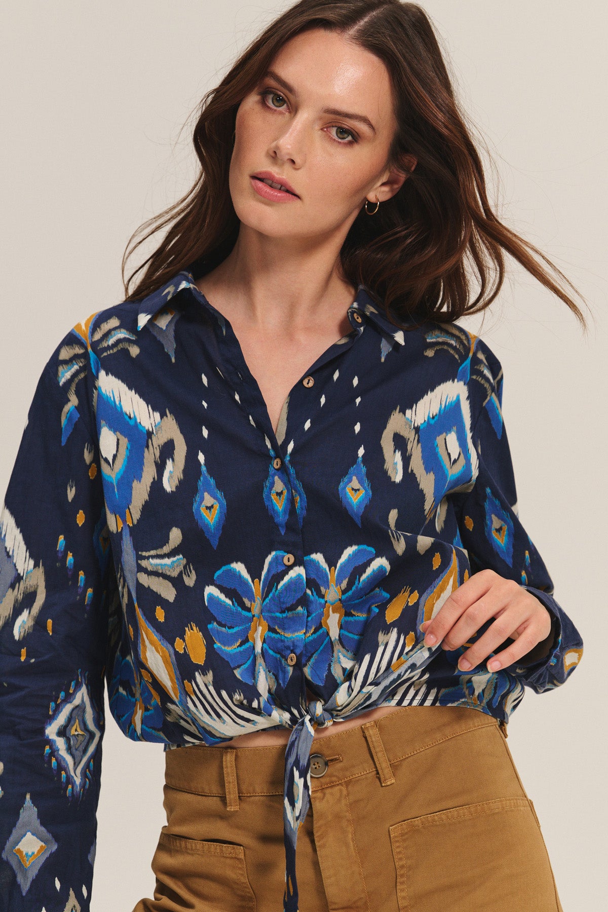 A woman models the KAYLA TOP by Velvet by Graham & Spencer, a blue patterned cotton-viscose blend button-down cropped blouse tied at the waist, paired with tan high-waisted pants against a plain background.-40672318357697