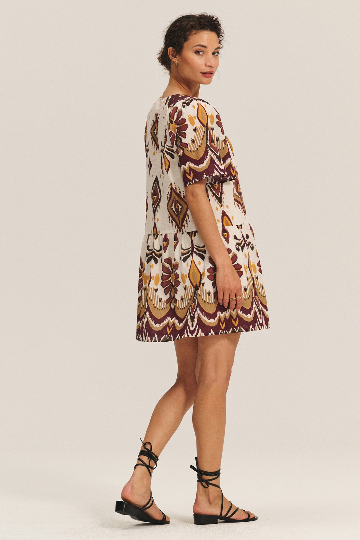 A woman stands sideways against a plain background, wearing the MAGGIE DRESS by Velvet by Graham & Spencer—a short-sleeve, patterned dress with an adjustable belt and white base featuring maroon and yellow designs—paired with black strappy sandals.-40598485500097
