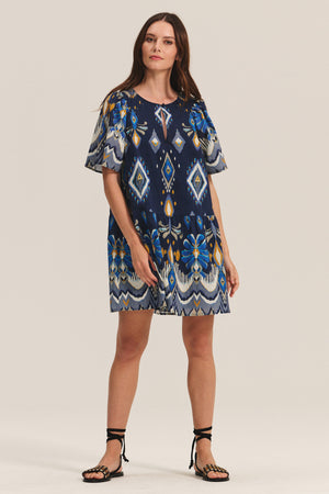 A woman wears the Velvet by Graham & Spencer MAGGIE DRESS, a blue patterned loose-fit dress with matching belt, styled with black lace-up sandals, standing against a plain background.