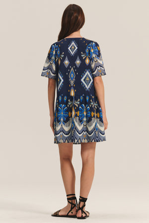 A person stands facing away, wearing the MAGGIE DRESS by Velvet by Graham & Spencer—a short-sleeved dress with blue, white, and yellow geometric patterns, a matching belt, and black sandals.