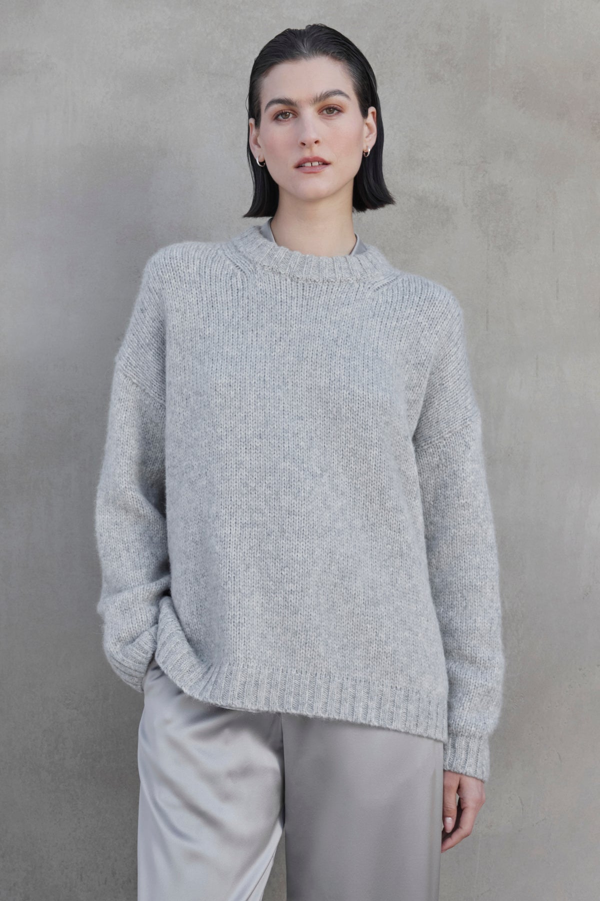   A person with short dark hair wears the CAMBRIA ALPACA SWEATER by Velvet by Jenny Graham, along with light gray pants, standing against a concrete wall. 