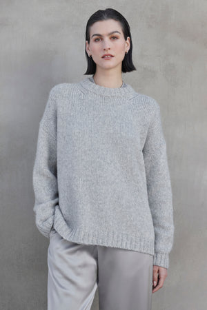 A person with short dark hair wears the CAMBRIA ALPACA SWEATER by Velvet by Jenny Graham, along with light gray pants, standing against a concrete wall.