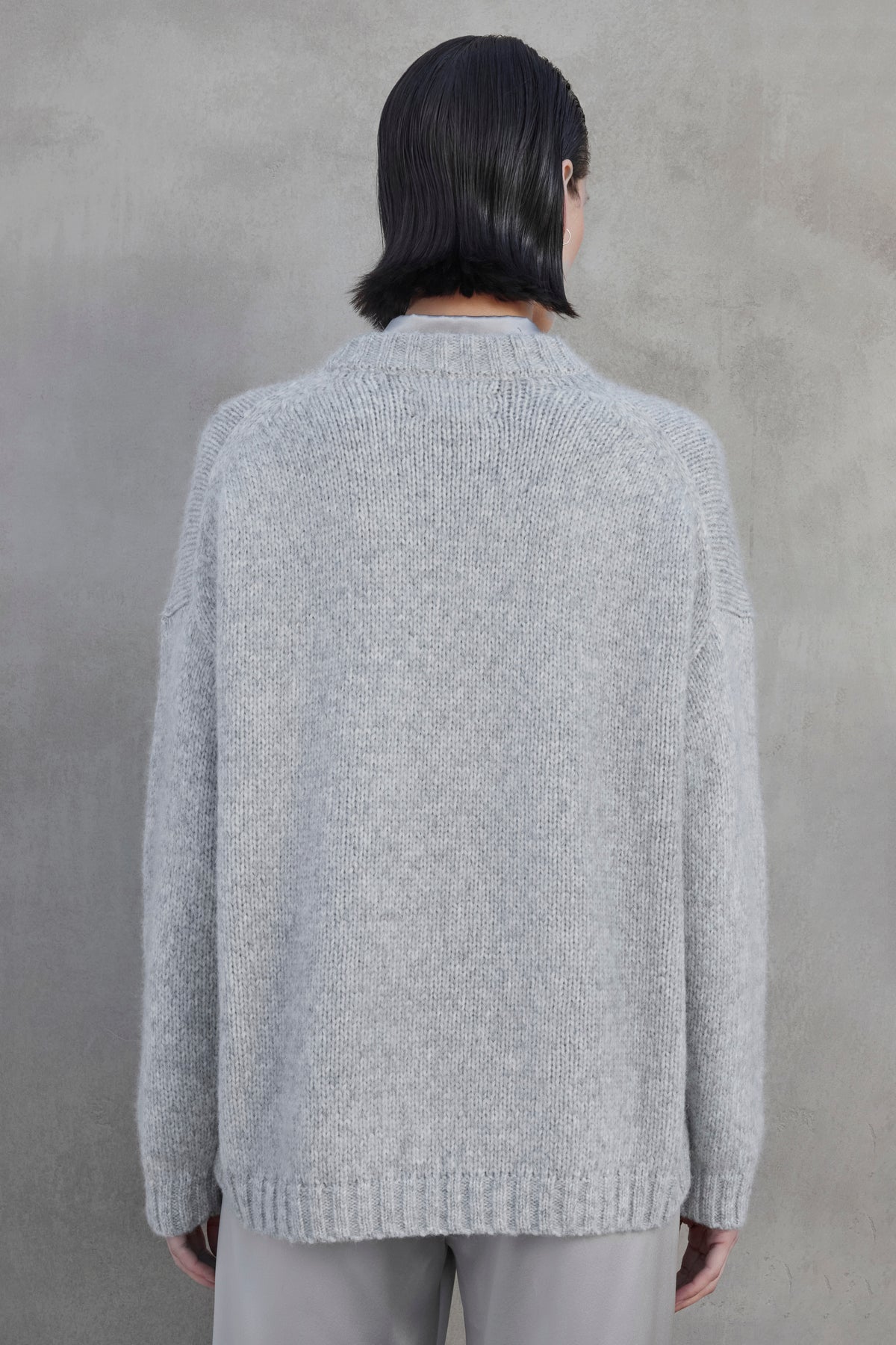 A person with short dark hair facing away, wearing an oversized CAMBRIA ALPACA SWEATER by Velvet by Jenny Graham and light-colored pants, standing against a plain gray wall.-37659958739137