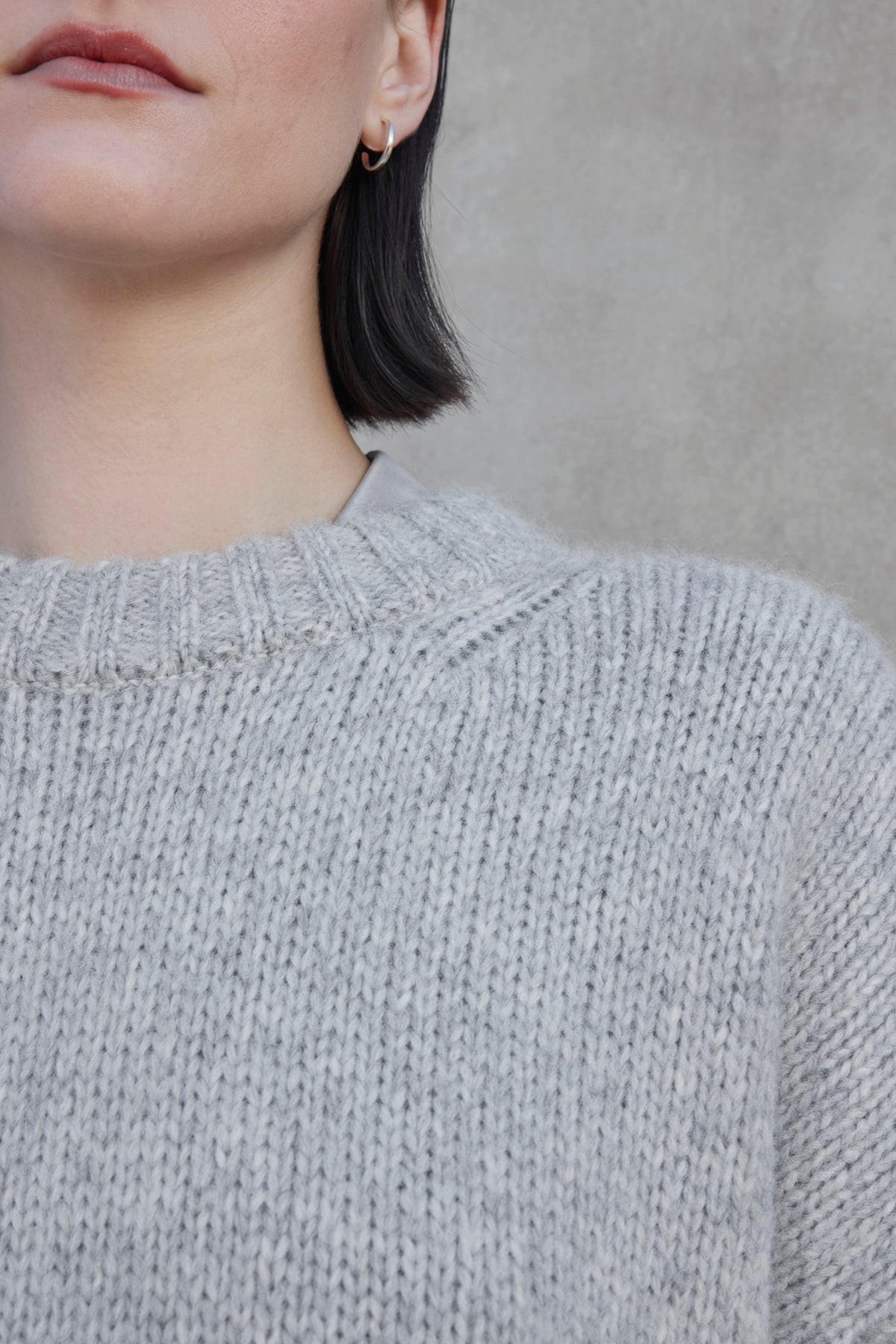   Close-up of a person wearing the CAMBRIA ALPACA SWEATER by Velvet by Jenny Graham in light gray, with a partial view of their face and short dark hair. The background is a plain, gray surface. 