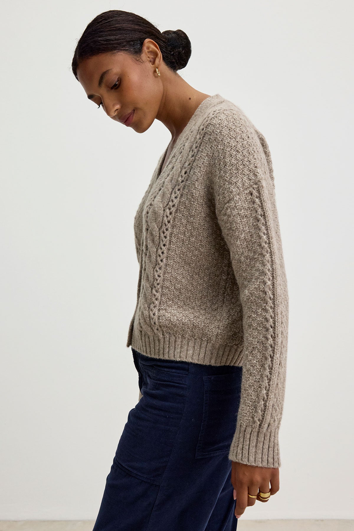   Person wearing the IZZY CARDIGAN by Velvet by Graham & Spencer, a beige alpaca blend with a four-button front, paired with dark blue pants, standing in profile against a plain background. 