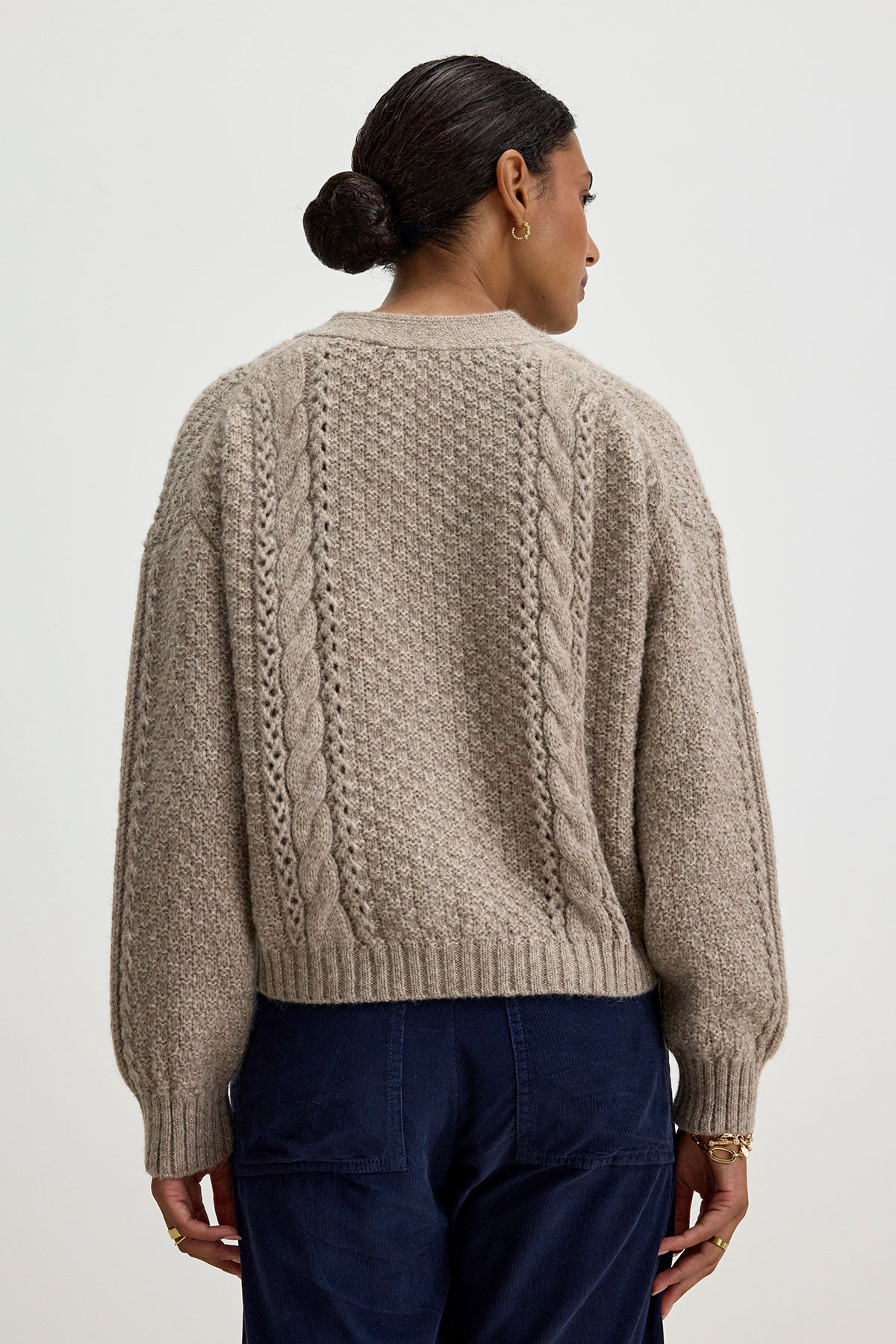   A person with dark hair in a bun is wearing the IZZY CARDIGAN by Velvet by Graham & Spencer along with dark blue pants, seen from the back. 