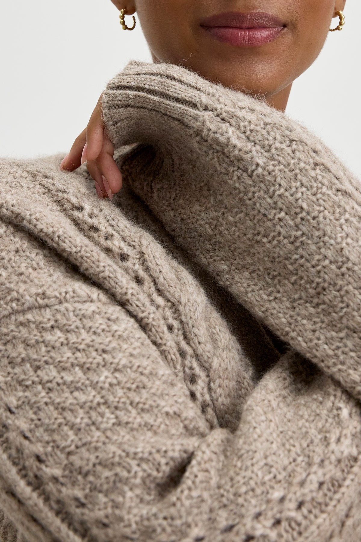   Close-up of a person wearing the IZZY CARDIGAN by Velvet by Graham & Spencer, featuring an alpaca blend with a textured beige, cable knit pattern, and one hand partially tucked into the sleeve. 