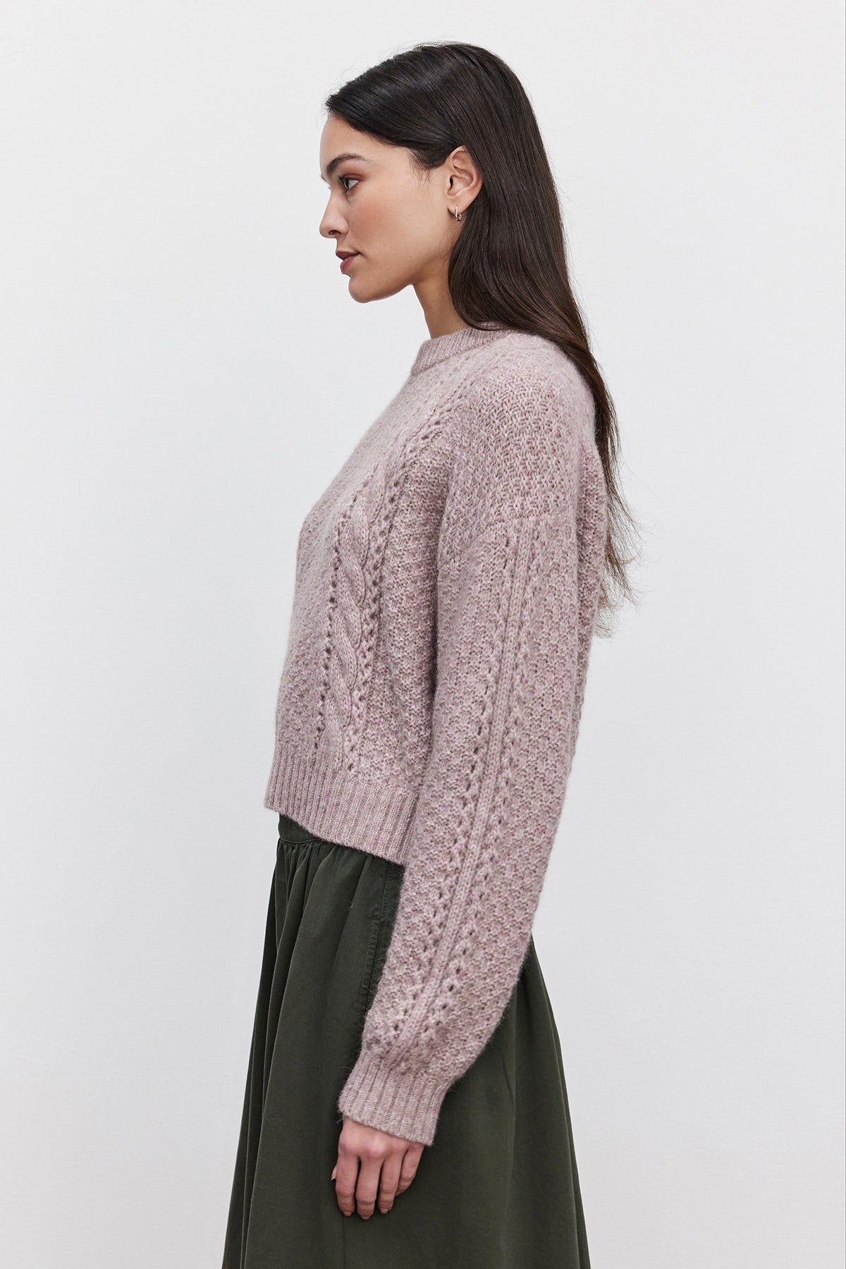   A person with long dark hair is wearing the MARLEY SWEATER from Velvet by Graham & Spencer, a light purple cable knit piece made from an alpaca blend, paired with an olive green skirt. They are standing in profile against a plain white background. 