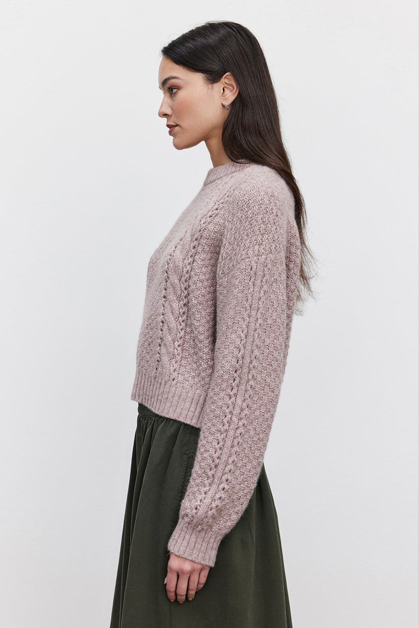 A person with long dark hair is wearing the MARLEY SWEATER from Velvet by Graham & Spencer, a light purple cable knit piece made from an alpaca blend, paired with an olive green skirt. They are standing in profile against a plain white background.