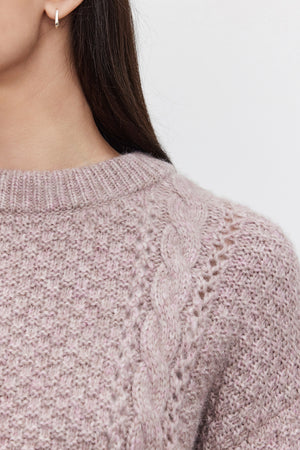 Close-up of a person wearing the MARLEY SWEATER by Velvet by Graham & Spencer, showcasing its light purple alpaca blend fabric with a braided pattern and accessorized with small hoop earrings, capturing only the shoulder and part of the face.