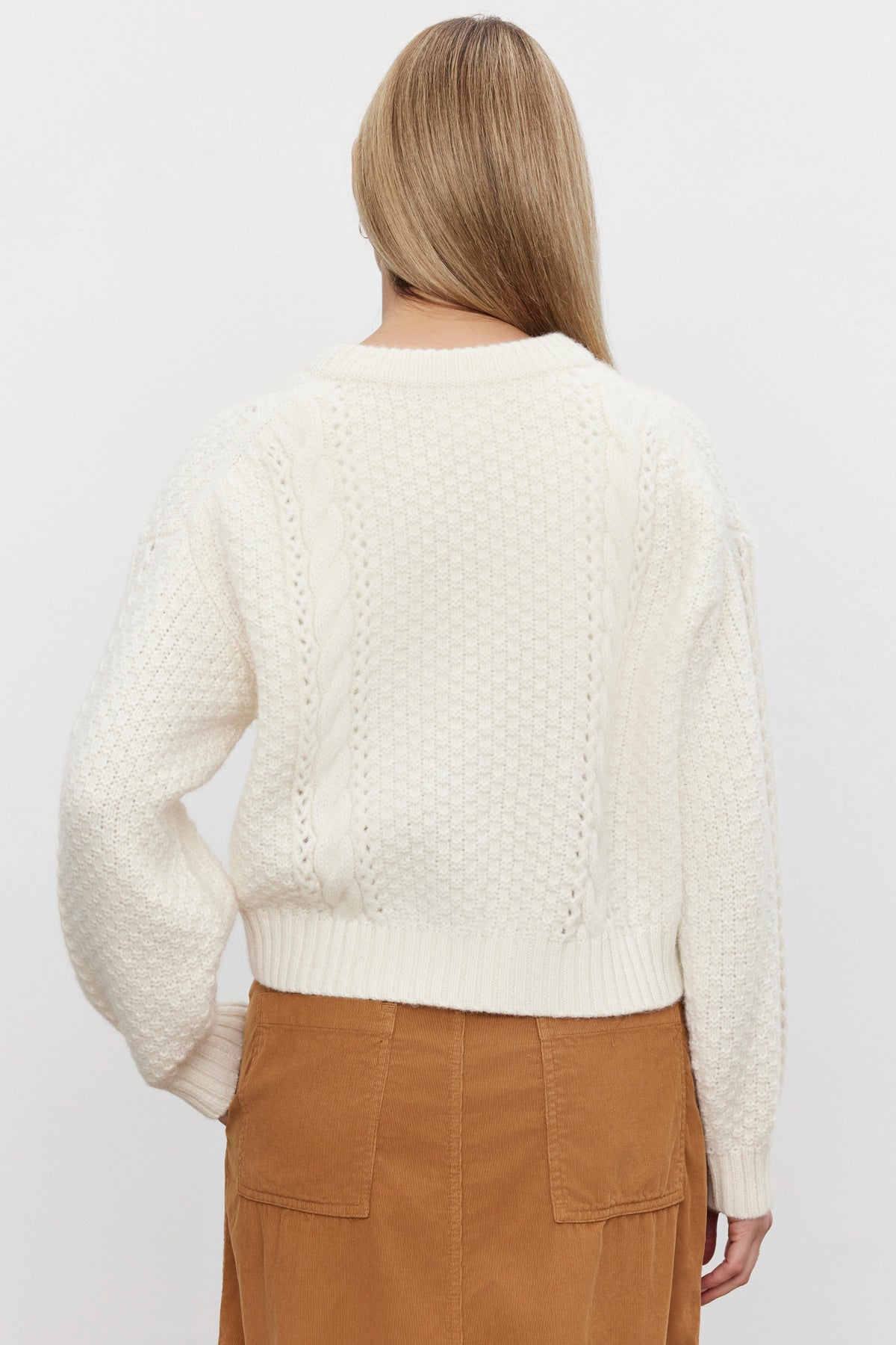   A person with long, light-colored hair is facing away from the camera, wearing the MARLEY SWEATER by Velvet by Graham & Spencer in white with a cable knit design and a brown skirt. 