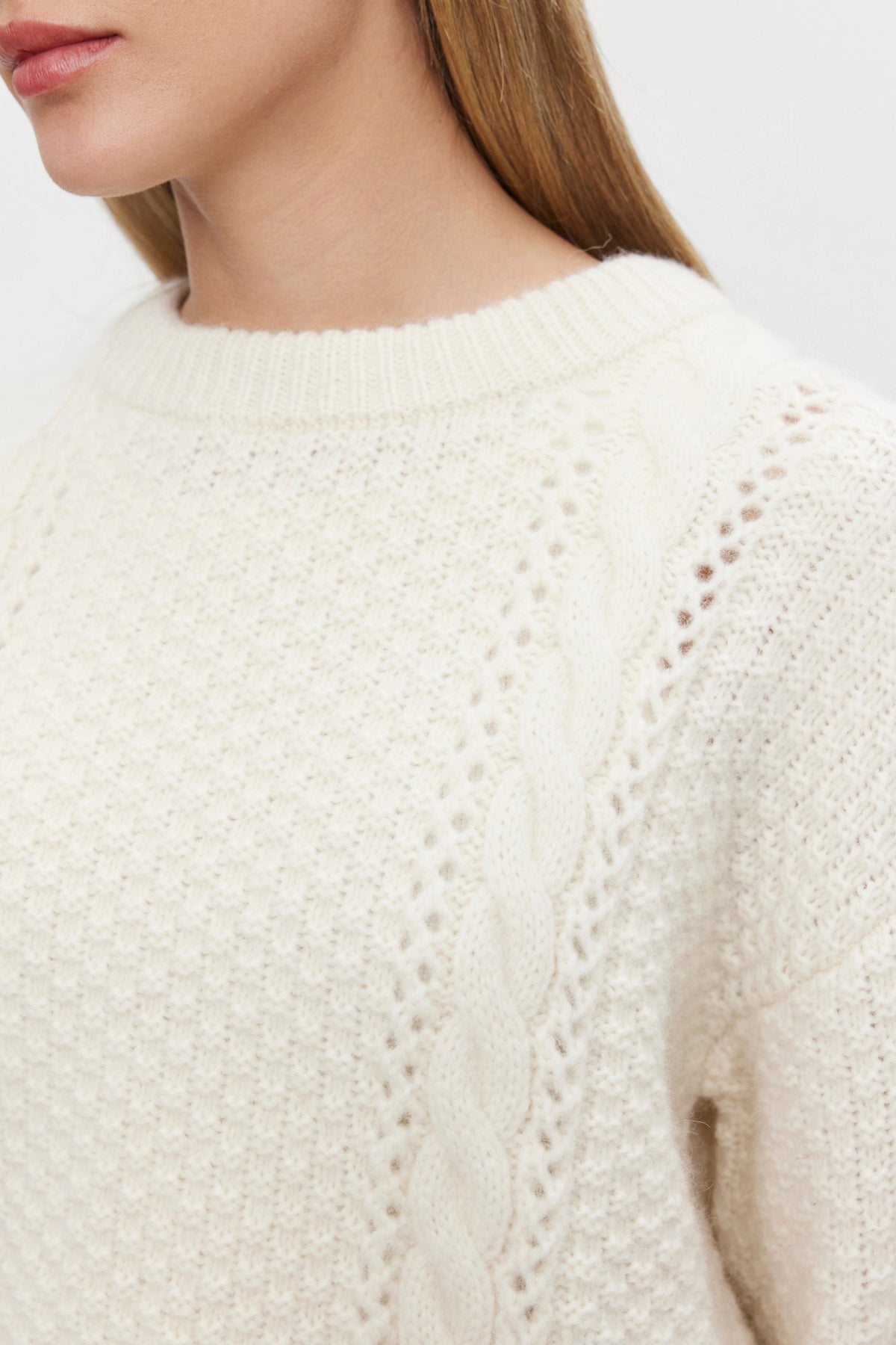   Close-up of a person wearing the MARLEY SWEATER by Velvet by Graham & Spencer, a cream-colored, cropped alpaca blend sweater with a vertically running cable-knit pattern on the shoulder. The person's long, blonde hair frames their face, of which only the lower half is visible. 