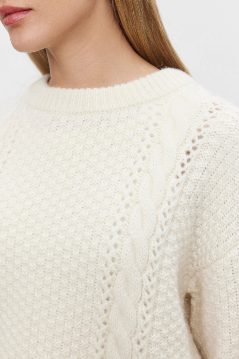 Close-up of a person wearing the MARLEY SWEATER by Velvet by Graham & Spencer, a cream-colored, cropped alpaca blend sweater with a vertically running cable-knit pattern on the shoulder. The person's long, blonde hair frames their face, of which only the lower half is visible.