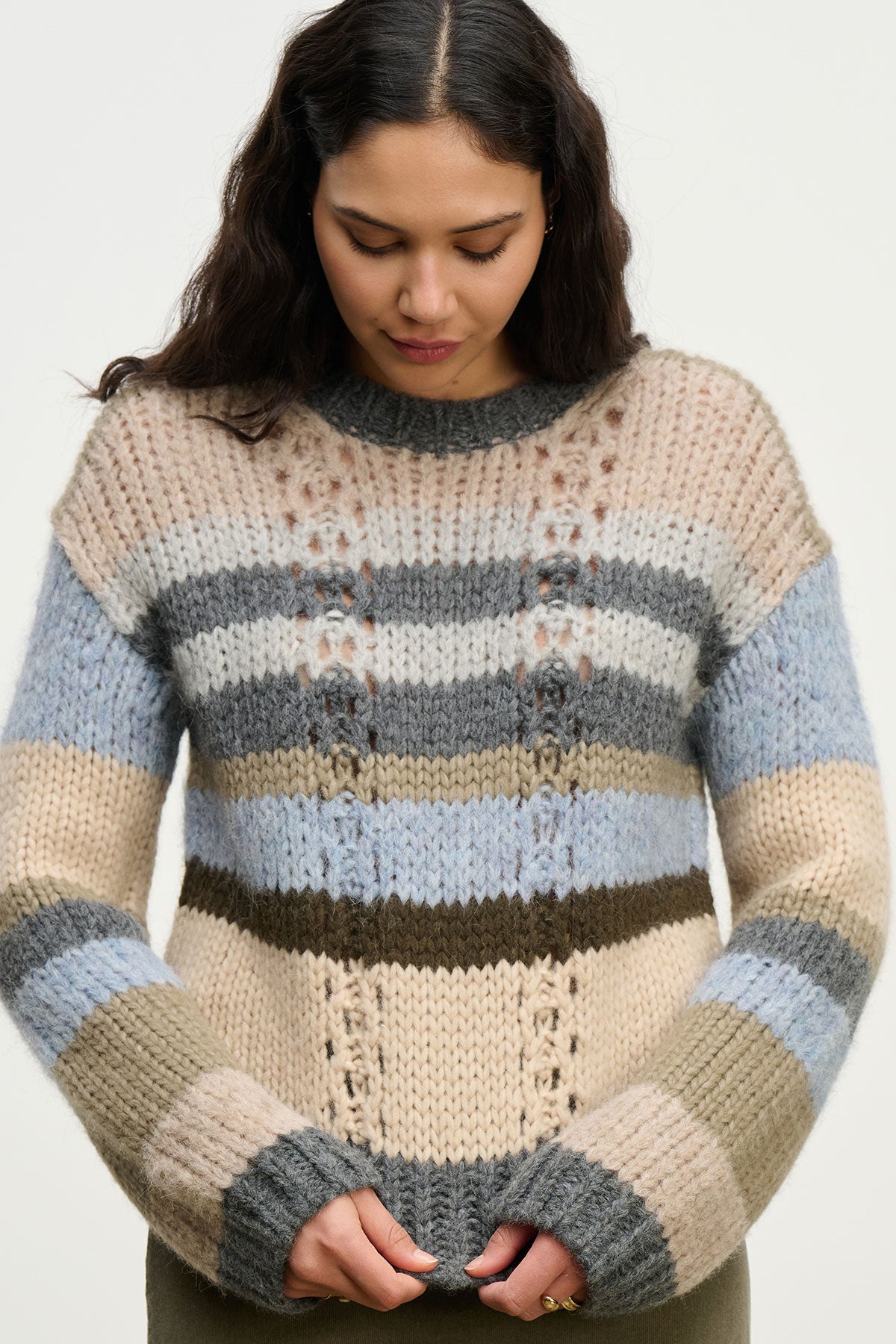A woman with long, dark hair looks down, wearing the RINA ALPACA SWEATER by Velvet by Graham & Spencer—a chunky alpaca blend in neutral and blue stripes with a boxy fit—set against a plain background.-40182795436225