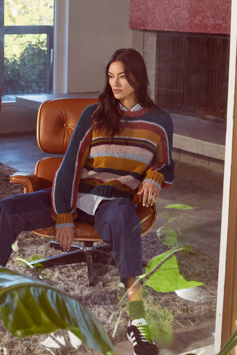 A woman sitting on a cozy chair in a living room wearing the Velvet by Graham & Spencer SAMARA STRIPED CREW NECK SWEATER.