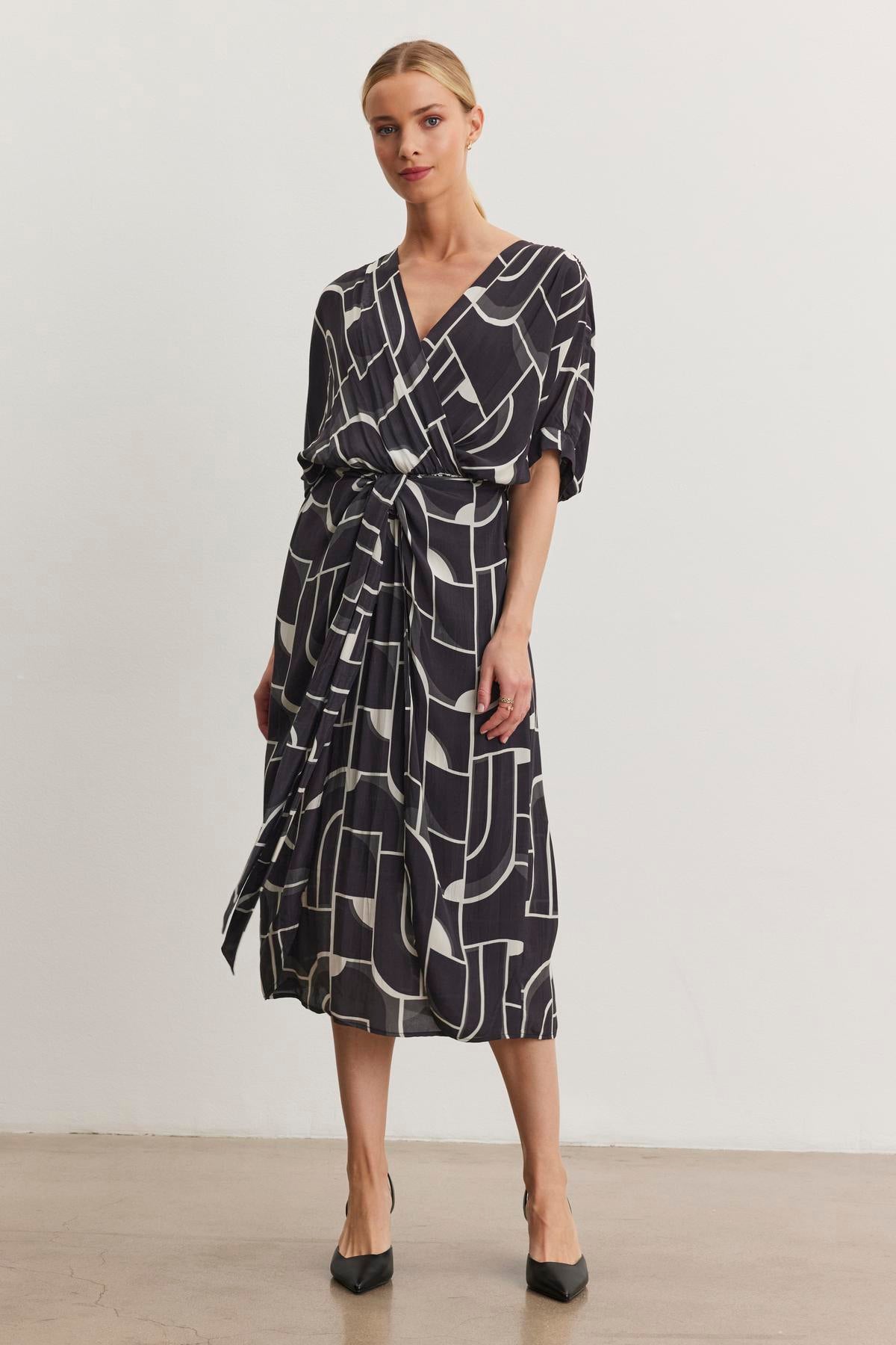   A person stands against a plain background wearing the ELISE DRESS by Velvet by Graham & Spencer, featuring Bauhaus-inspired prints with wrap front ties, paired with black heels. 