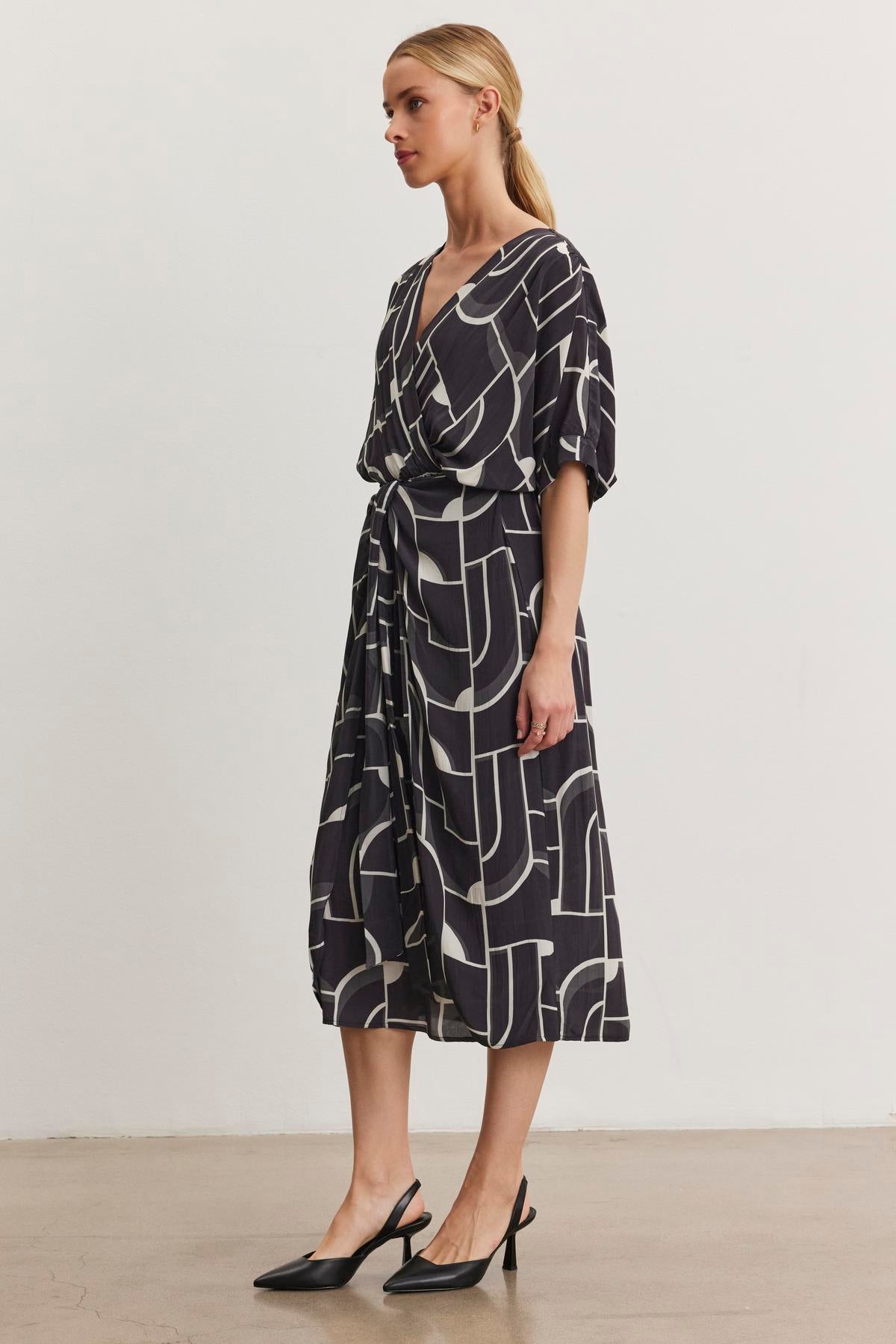 A person stands sideways in the ELISE DRESS by Velvet by Graham & Spencer, a black and white V-neck dress with Bauhaus-inspired print, featuring short sleeves and paired with black heels.-38512546513089