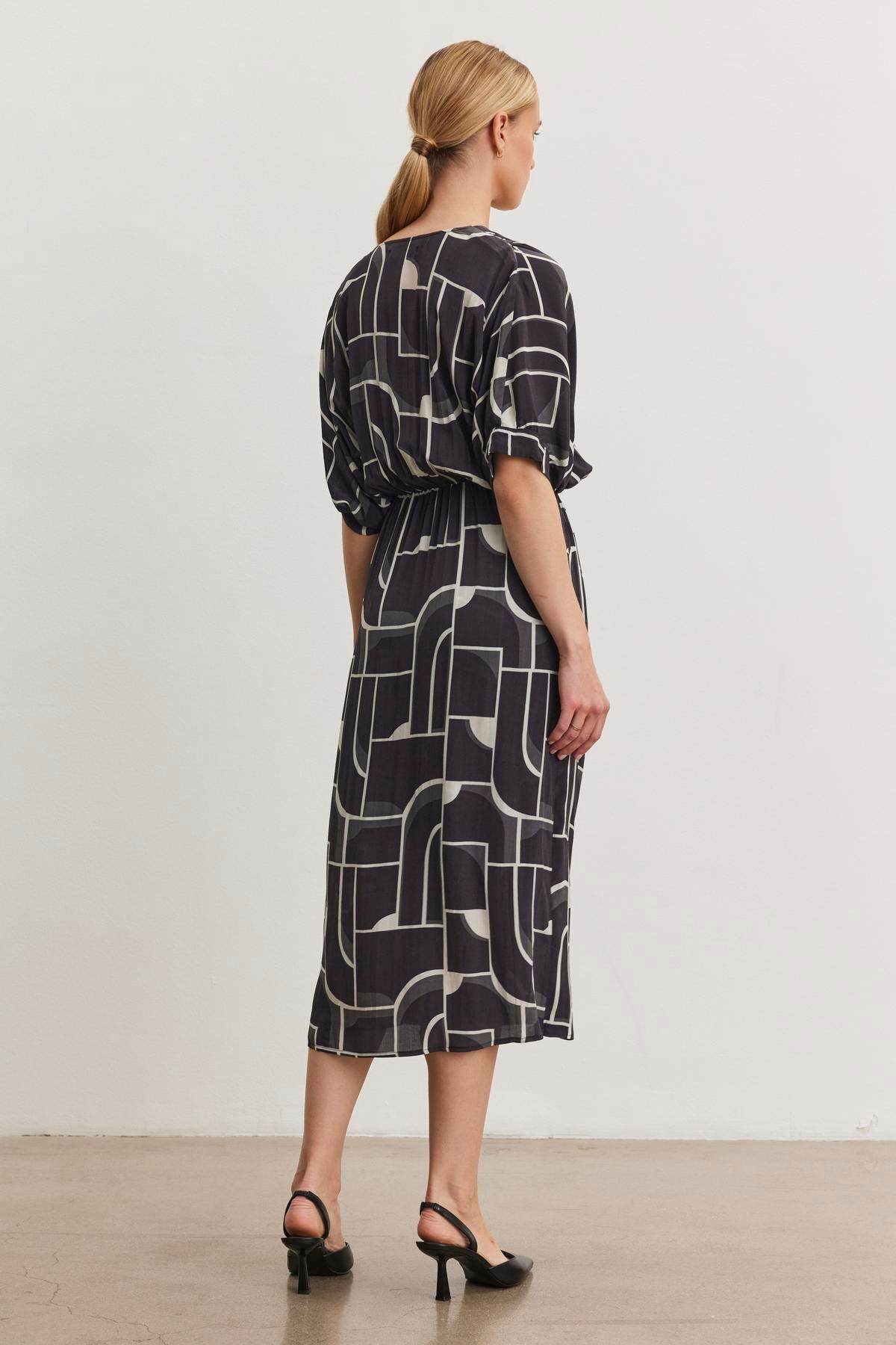 A blonde woman stands wearing the ELISE DRESS by Velvet by Graham & Spencer, a black dress with a Bauhaus-inspired geometric pattern. The V-neckline wrap-front dress is paired with elegant black heels as she faces a plain white wall.-38512546545857