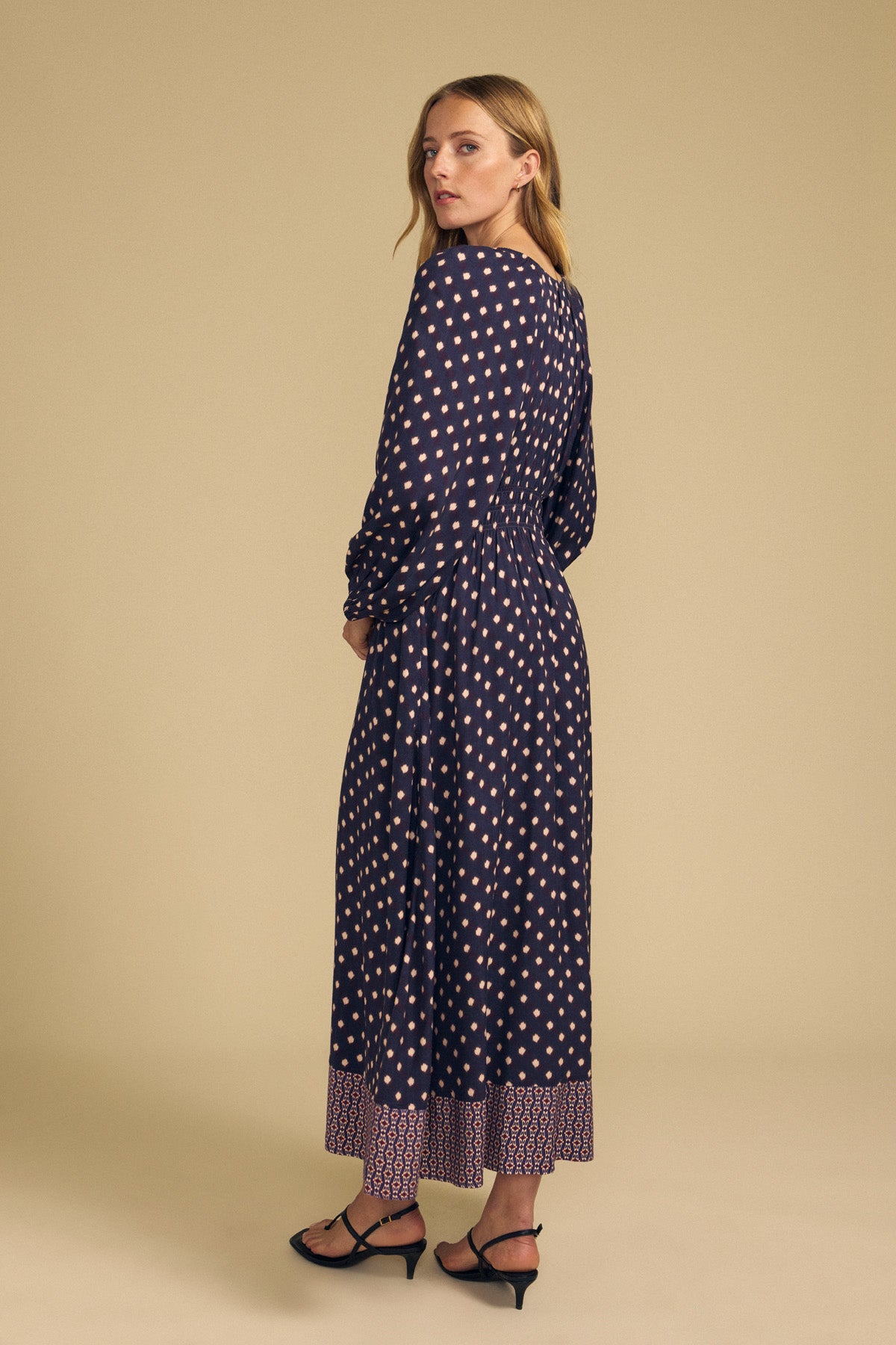 A woman wearing a navy blue MALLORY PRINTED DRESS with a diamond pattern and border print, paired with black strappy low-heeled sandals for a special occasion.-46489566183617