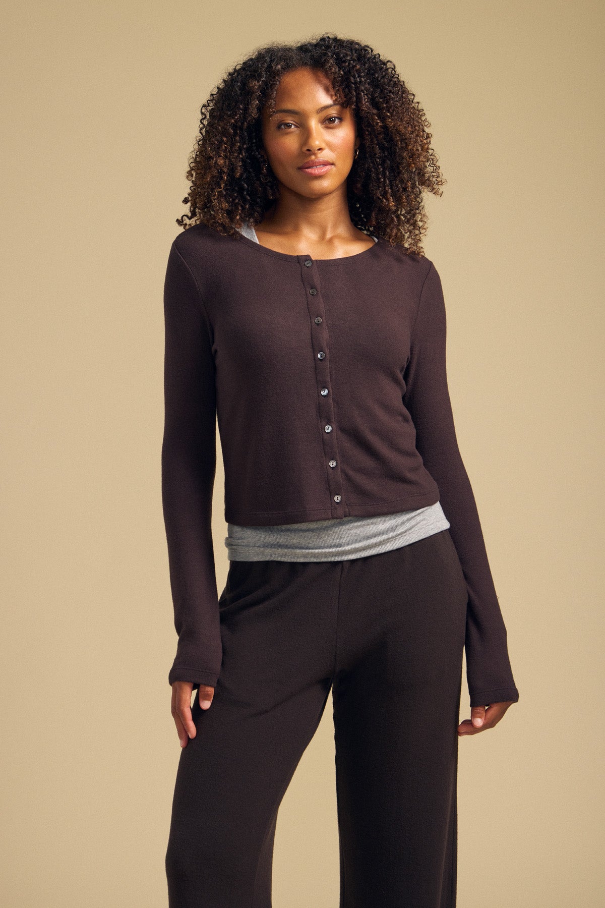 A woman wears the BEATRICE CARDIGAN by Velvet by Graham & Spencer in brown, buttoned up over a gray top and matching brown pants, standing against a beige background.-42063723954369