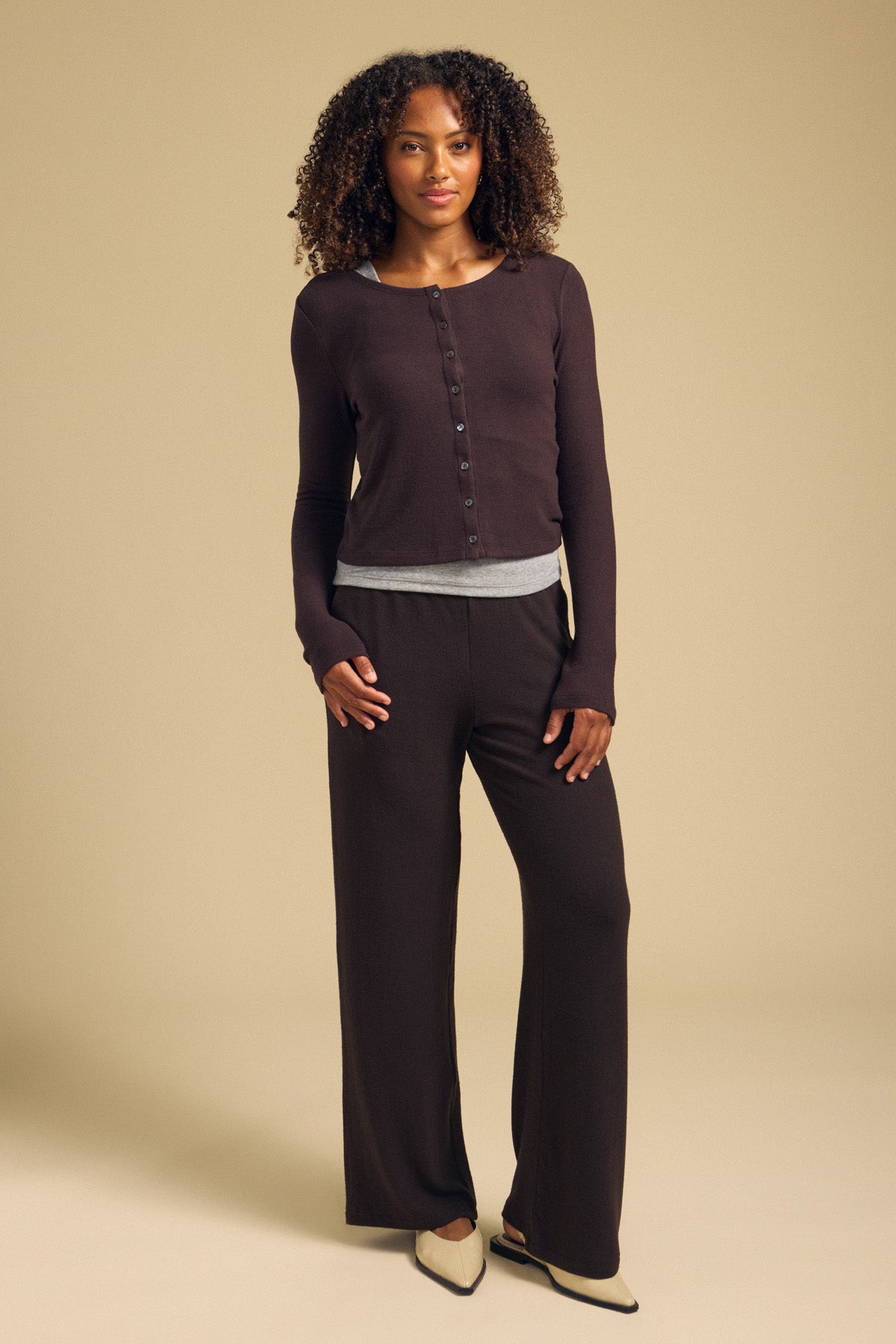 A woman stands against a beige background wearing the BEATRICE CARDIGAN by Velvet by Graham & Spencer, layered over a gray undershirt, paired with matching wide-leg pants and cream-colored pointed flats.