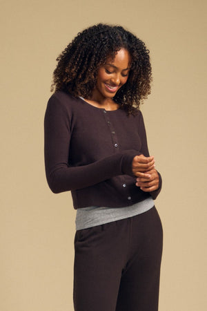 Wearing the BEATRICE CARDIGAN by Velvet by Graham & Spencer, a woman with curly hair stands against a beige background, smiling as she looks down. The soft knit, dark brown buttoned cardigan is layered over a gray shirt and matching brown pants for an effortlessly stylish look.