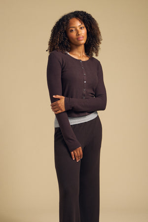 A woman stands against a beige background, wearing the BEATRICE CARDIGAN by Velvet by Graham & Spencer—a dark brown cropped cardigan—layered over matching pants, with a gray shirt visible at the waist.