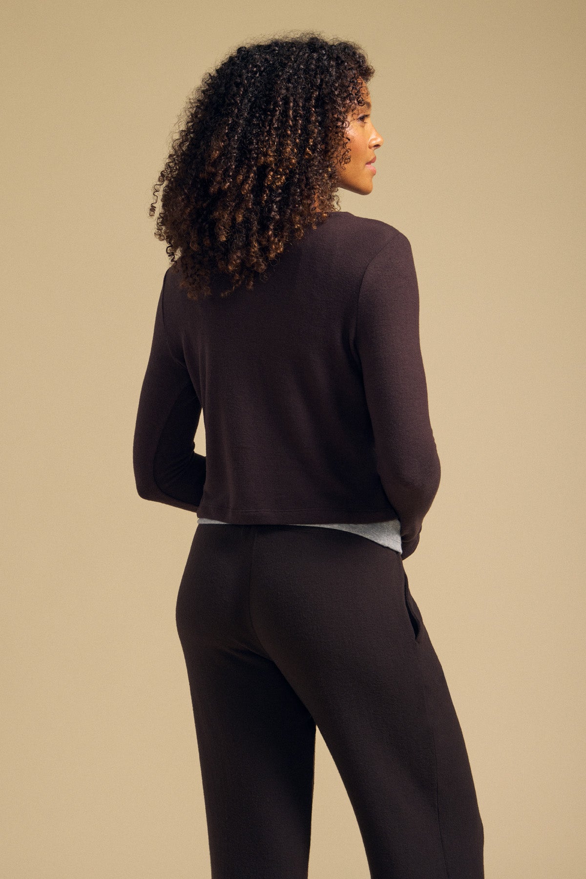 A woman with curly hair faces away, wearing the BEATRICE CARDIGAN by Velvet by Graham & Spencer with matching pants, set against a plain beige background.