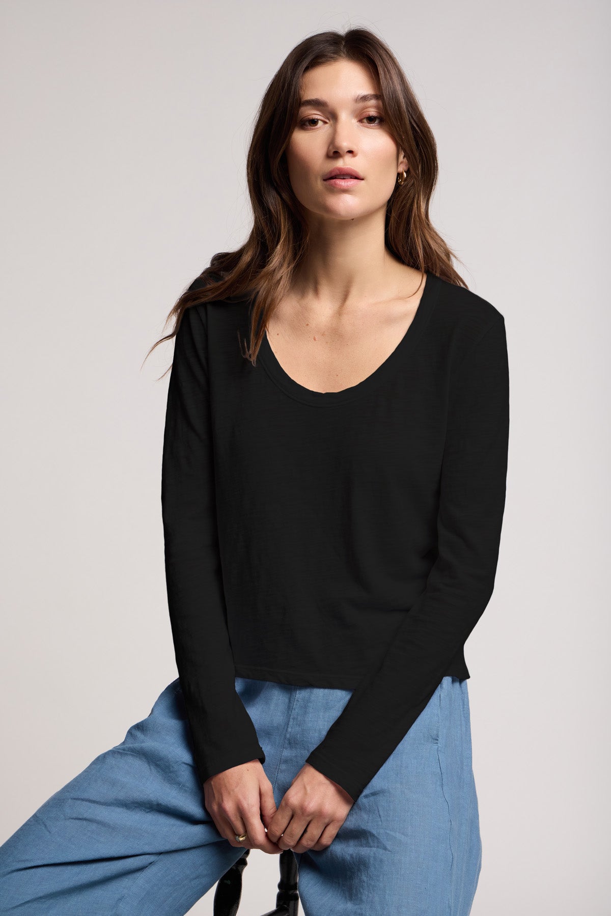 A woman with long brown hair sits on a stool against a plain background, wearing the Velvet by Graham & Spencer BECKY TEE—a black cotton slub long-sleeve top—paired with light blue pants.
