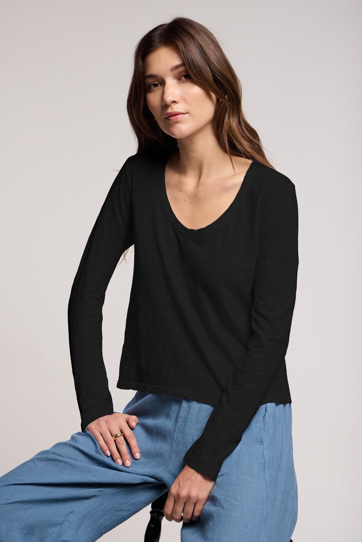 A woman with long brown hair sits on a stool against a plain background, wearing the Velvet by Graham & Spencer BECKY TEE—a relaxed scoop neck black cotton slub long-sleeve top—with light blue pants.-41014994403521