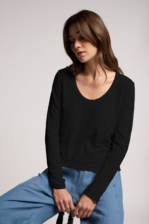 A woman with long brown hair wears the Velvet by Graham & Spencer BECKY TEE, a black relaxed scoop neck long-sleeve, with light blue pants while sitting against a plain background.