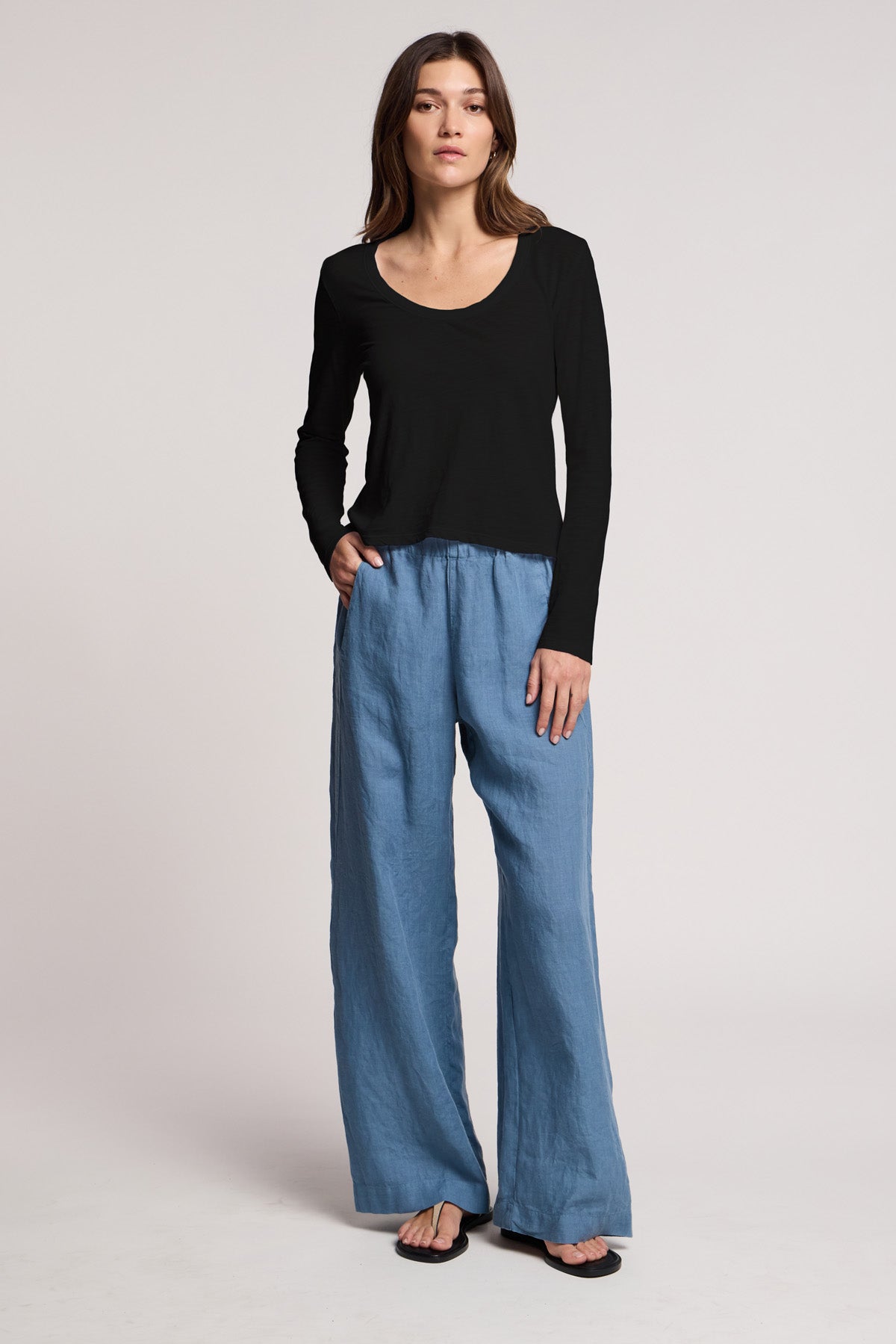 A woman stands against a neutral background wearing the BECKY TEE by Velvet by Graham & Spencer, a relaxed scoop neck black cotton slub long-sleeve tee, paired with loose blue pants and black sandals, one hand in her pocket.-41014994469057