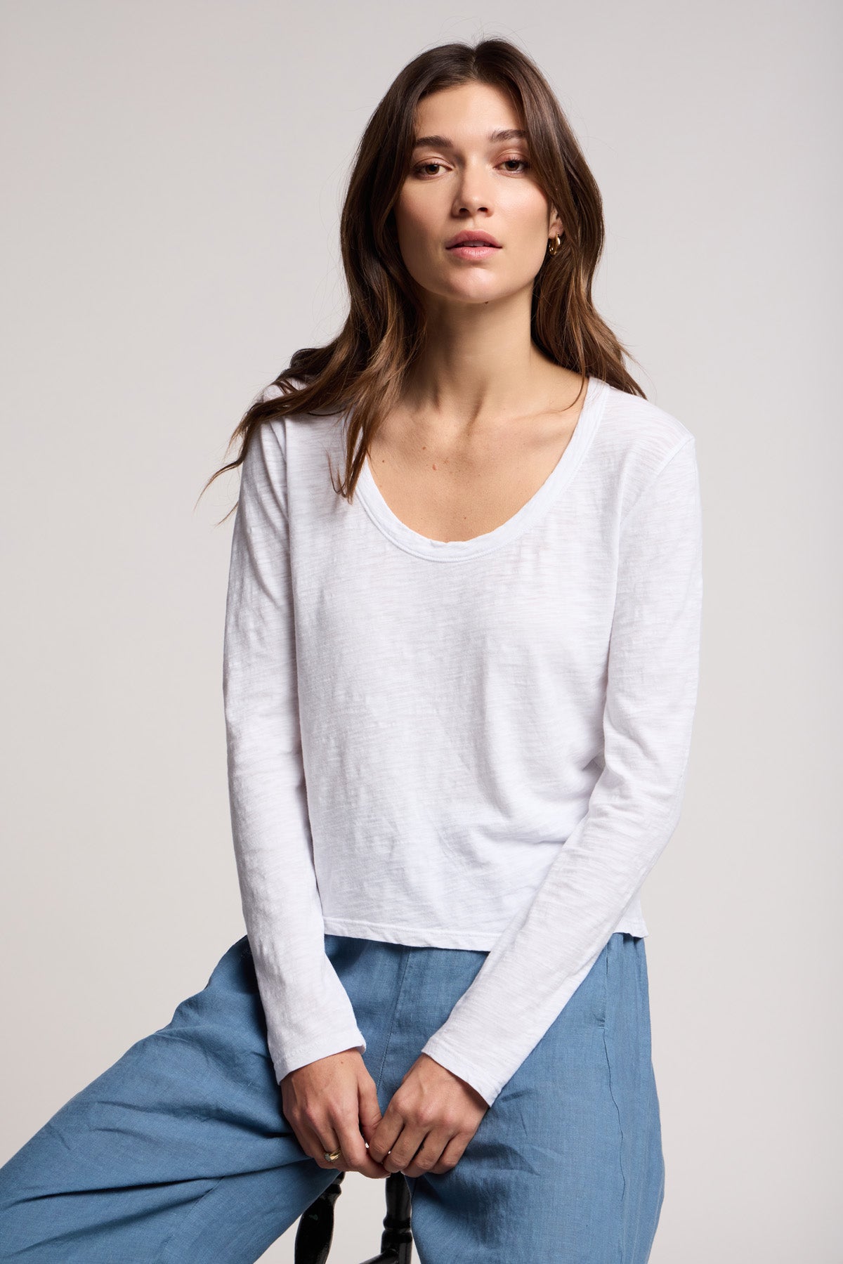 A woman with long brown hair wears the BECKY TEE by Velvet by Graham & Spencer, a relaxed scoop neck long-sleeve top, paired with light blue pants, seated against a plain light background.-40475878228161