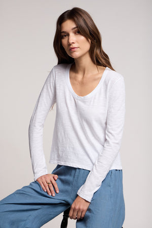 A woman with long brown hair wears the BECKY TEE by Velvet by Graham & Spencer—a white cotton slub top with a relaxed scoop neck—paired with light blue pants, seated on a stool against a plain light background.