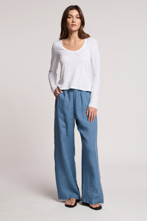 A woman stands against a plain background wearing the BECKY TEE by Velvet by Graham & Spencer—a white, relaxed scoop neck long-sleeve top—paired with blue wide-leg pants and black sandals, one hand in her pocket.
