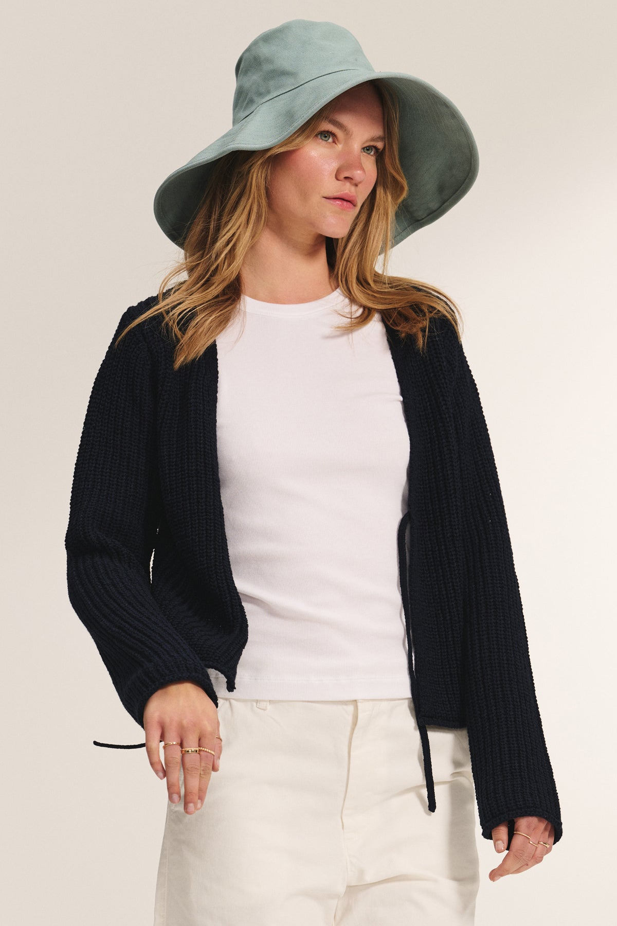 A woman stands against a neutral background wearing a light blue wide-brim hat, black fitted cardigan, white pants, and the BEDFORD TEE by Velvet by Jenny Graham.-39172744085697