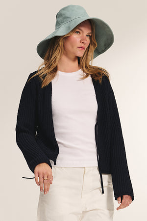 A woman wearing a light blue wide-brim hat, white t-shirt, cream pants, and the THALIA CARDIGAN by Velvet by Graham & Spencer stands against a plain background.